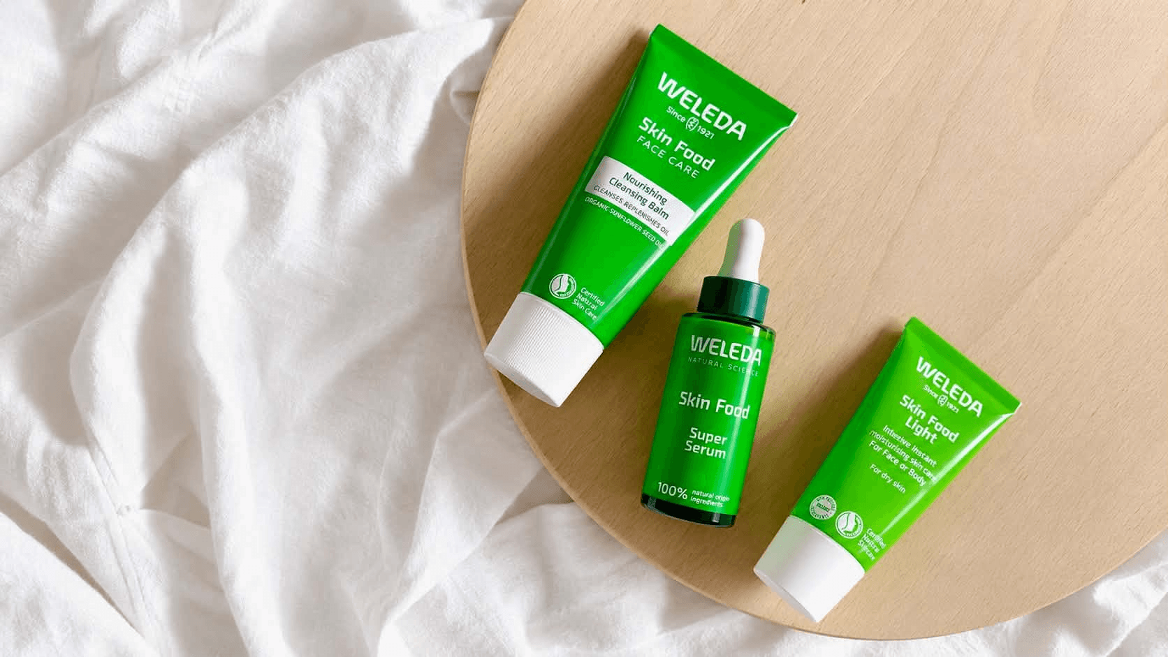 Balsam do mycia twarzy Weleda Skin Food, Super Serum i krem Skin Food Light na drewnianej tacy obok białej tkaniny